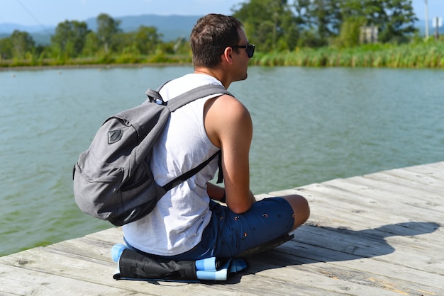 休んで池の桟橋に座っているバックパックを持つ若い男