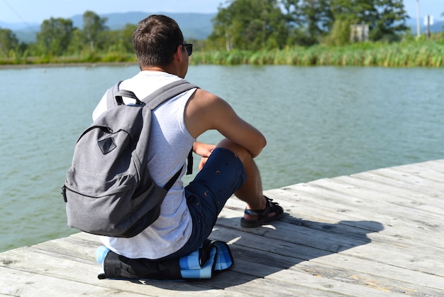 休んで池の桟橋に座っているバックパックを持つ若い男