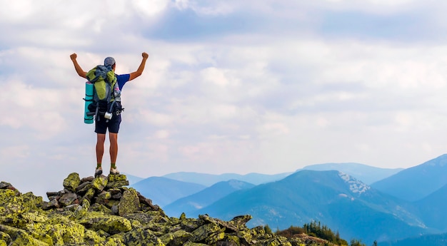 Young man with backpack standing with raised hands on top of a mountain and enjoying mountain view.