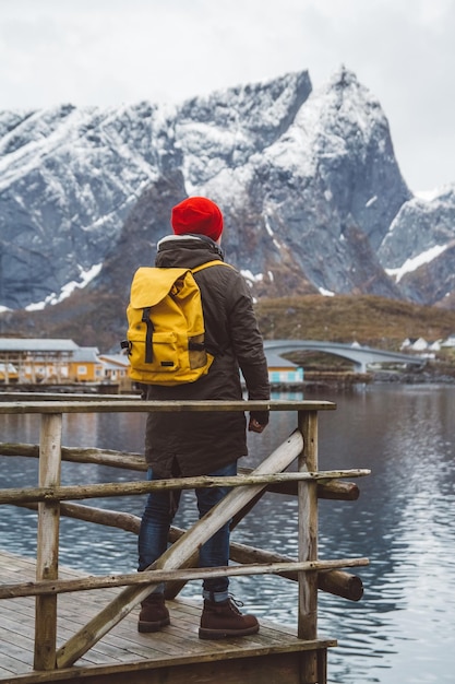 Young man with a backpack standing on a wooden pier the background of snowy mountains and lake