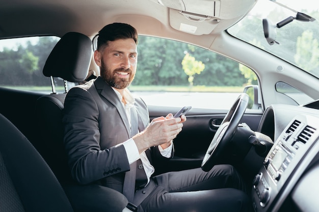 駐車場で車を運転しているひげのビジネスマンと若い男は笑顔で電話に書き込み、成功した男性のニュースを読みます