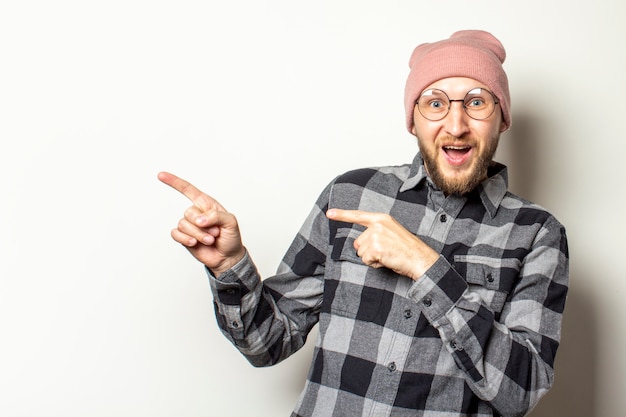 young man with a beard in a hat, a plaid shirt and glasses points his fingers to the side with a surprised face on an isolated white. Gesture look at it