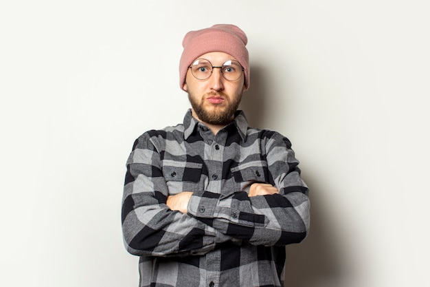 young man with a beard in a hat, plaid shirt and glasses with arms crossed on his chest and a serious face on an isolated white.