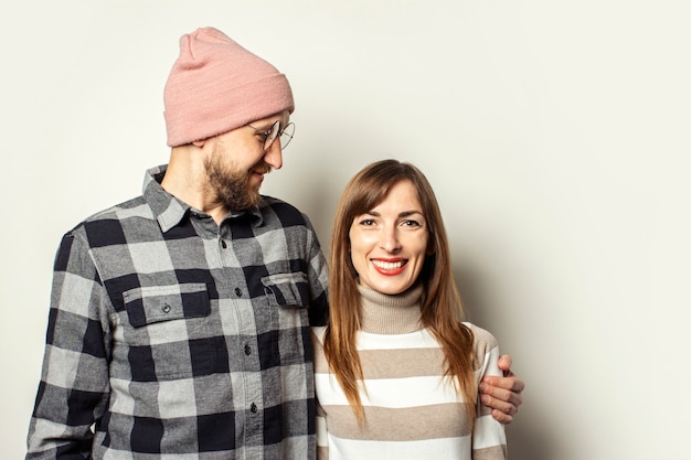 Young man with a beard in a hat and a plaid shirt hugs a girl in a sweater on an isolated light background.