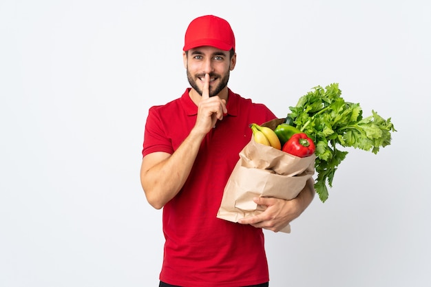 沈黙のジェスチャーをしている白い背景で隔離の野菜でいっぱいのバッグを保持しているひげを持つ若い男
