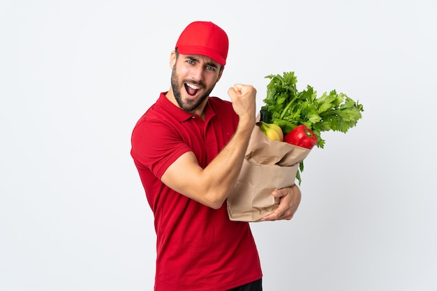 強いジェスチャーを作る白い背景で隔離の野菜でいっぱいのバッグを保持しているひげを持つ若い男