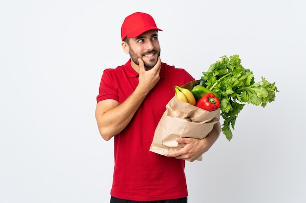 アイデアを考えて白で隔離される野菜の完全な袋を保持しているひげを持つ若者