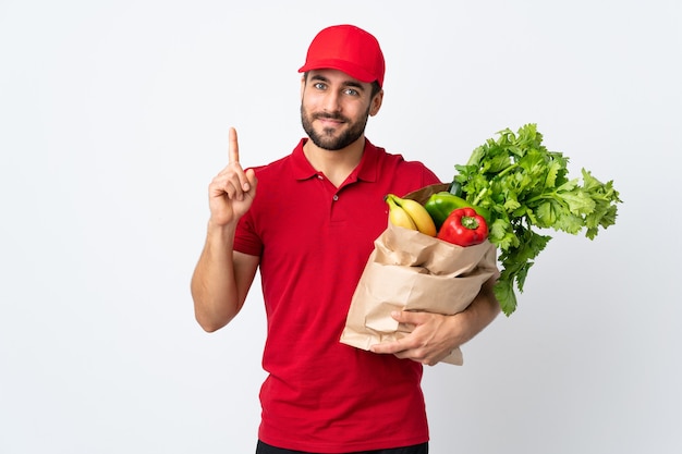 人差し指で指している白い壁に分離された野菜でいっぱいのバッグを保持しているひげを持つ若い男は素晴らしいアイデア