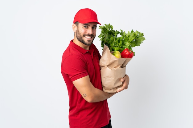 Молодой человек с бородой, держа сумку с овощами на белой стене смеется