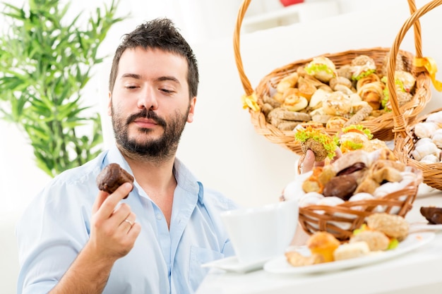 수염을 기른 청년, 손에 달콤하고 맛있는 패스트리를 들고 있습니다. 맛있는 음식을 바라보는 미소로. 배경에서 패스트리와 함께 짠 바구니를 볼 수 있습니다.