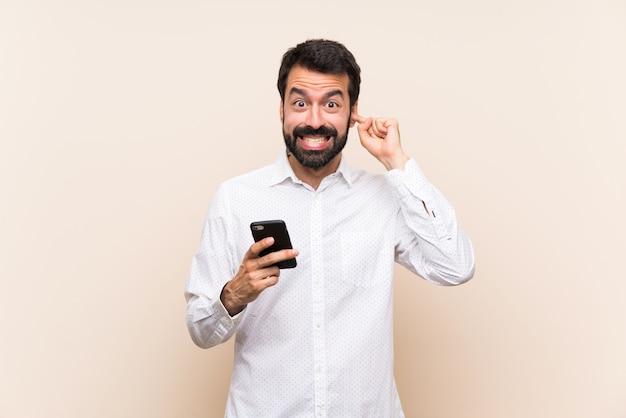 Young man with beard holding a mobile frustrated and covering ears