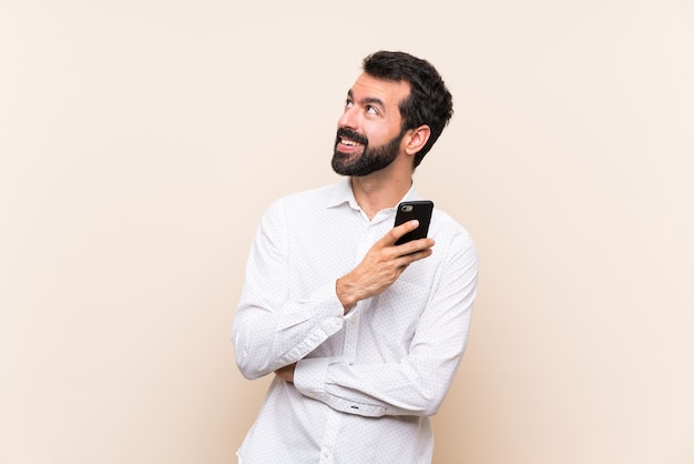 Young man with beard holding a mobile looking up while smiling