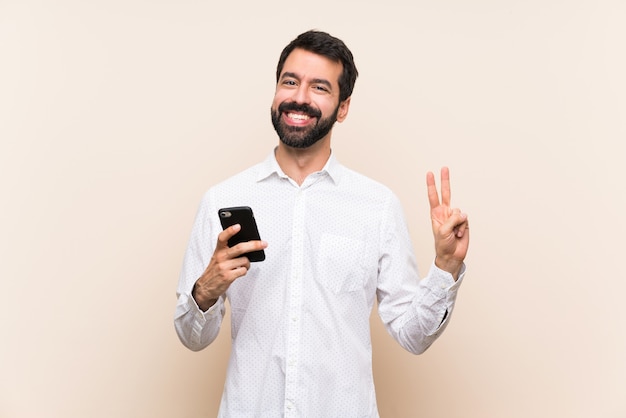 Young man with beard holding a mobile showing victory sign with both hands