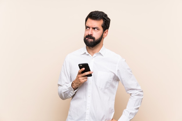 Молодой человек с бородой держит мобильный телефон с выражением лица путать