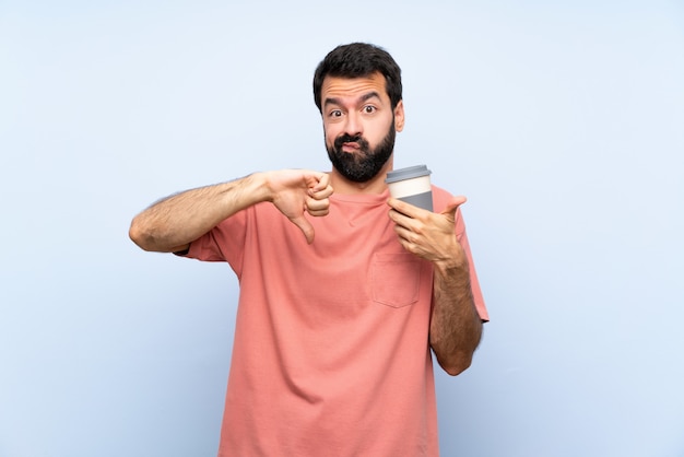 Young man with beard holding a take away coffee over isolated blue  making good-bad sign. Undecided between yes or not