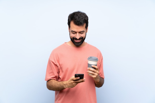 Young man with beard holding a take away coffee over isolated blue sending a message with the mobile