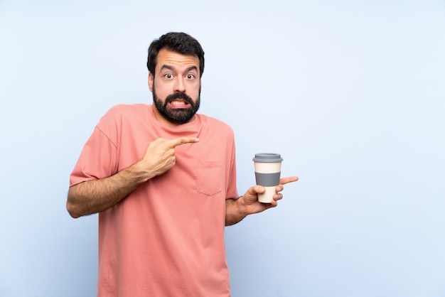 Young man with beard holding a take away coffee over isolated blue wall frightened and pointing to the side