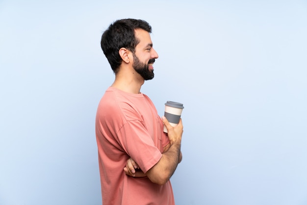 Young man with beard holding a take away coffee over isolated blue wall in lateral position