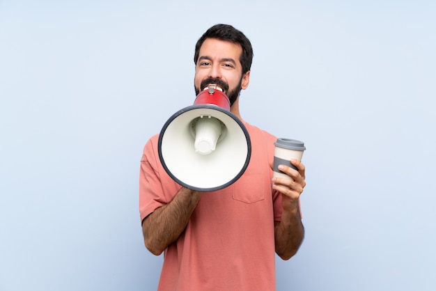 Young man with beard holding a take away coffee over isolated blue wall shouting through a megaphone