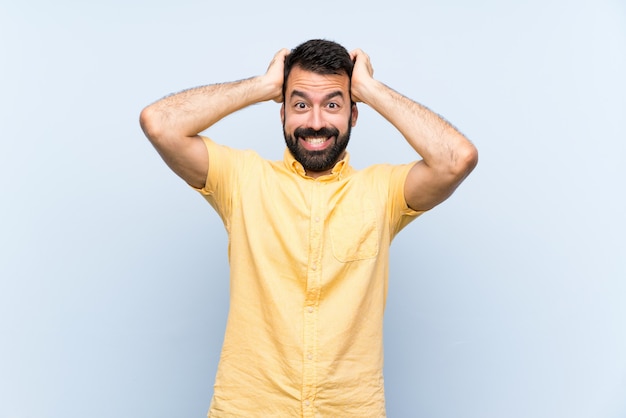 Young man with beard over isolated blue wall frustrated and takes hands on head