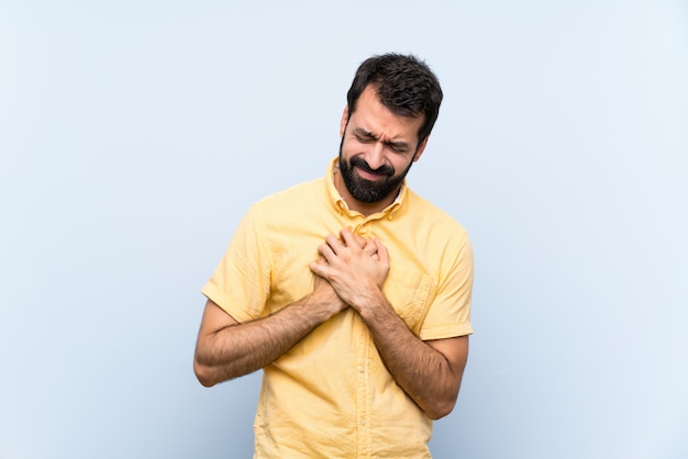 Young man with beard over isolated blue wall having a pain in the heart