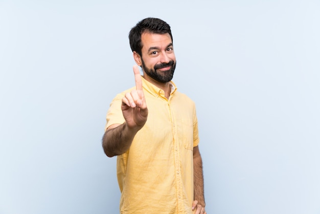 Young man with beard over isolated blue wall showing and lifting a finger