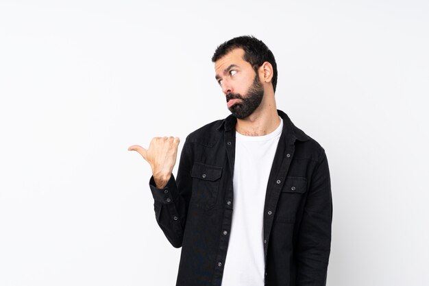 Young man with beard over isolated white  unhappy and pointing to the side