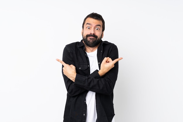 Young man with beard over isolated white wall pointing to the laterals having doubts