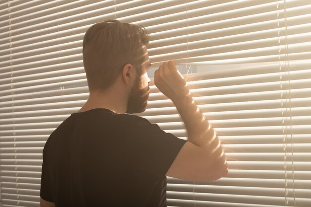 Young man with beard peeking through window blinds