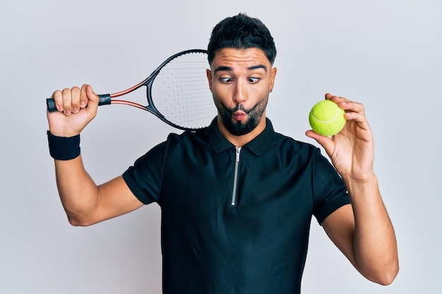 Молодой человек с бородой играет в теннис, держа ракетку и мяч, делая рыбье лицо со ртом и щурясь, безумно и комично