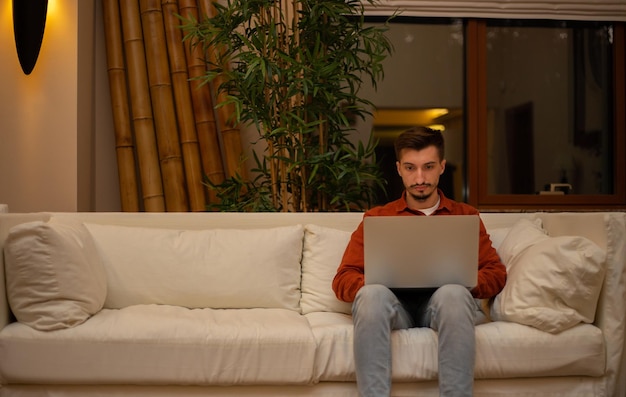 Photo a young man with a beard in a red shirt works with a laptop and sits on the couch in the evening in the house.