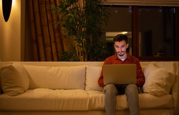 A young man with a beard in a red shirt works with a laptop and sits on the couch in the evening in the house.