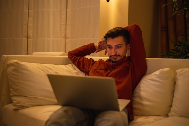 A young man with a beard in a red shirt works with a laptop and sits on the couch in the evening in the house.