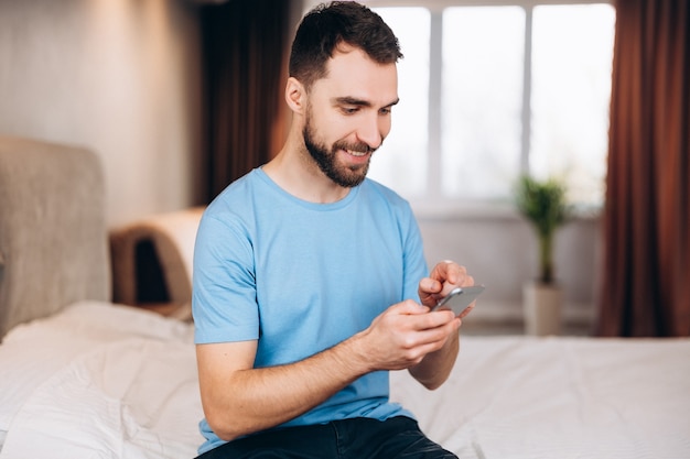 Young man with beard smiling whilst received sending message on phone while sitting in bed