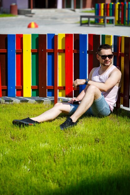 A young man with a beard in a T-shirt with a phone sits near a fence on a green lawn. Guy in sunglasses