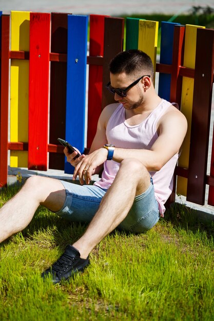 A young man with a beard in a T-shirt with a phone sits near a fence on a green lawn. Guy in sunglasses