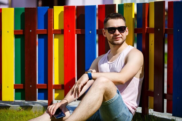 Photo a young man with a beard in a t-shirt with a phone sits near a fence on a green lawn. guy in sunglasses