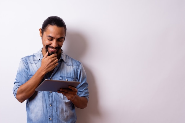 young man with beard tied hair smiles happy with hand on beard looks at his tablet and leaves the right side blank.