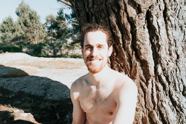 Young man with a beard next to a tree shirtless smiling to camera during a sunny day, confidence and holiday concept, portrait