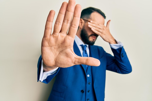 Young man with beard wearing business suit and tie covering eyes with hands and doing stop gesture with sad and fear expression. embarrassed and negative concept.