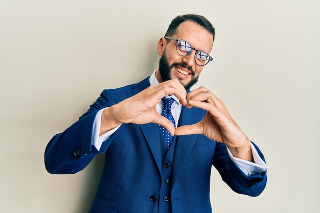 Young man with beard wearing business suit and tie smiling in love doing heart symbol shape with hands romantic concept