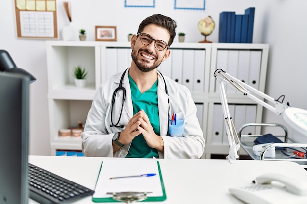 Photo young man with beard wearing doctor uniform and stethoscope at the clinic hands together and fingers crossed smiling relaxed and cheerful success and optimistic