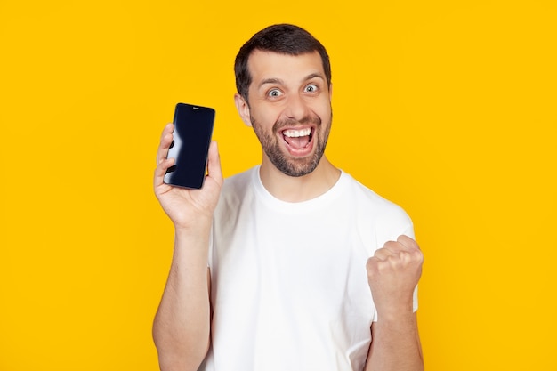 young man with a beard in a white T-shirt shows a smartphone screen, shouts with pride and celebrates victory and success