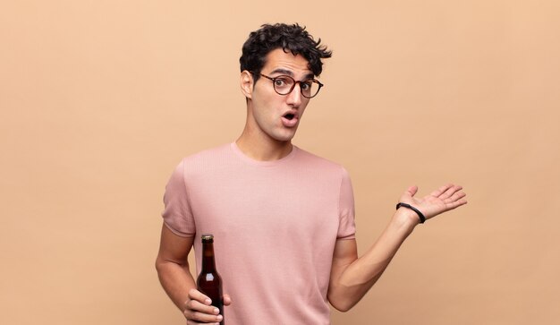 Young man with a beer looking surprised and shocked, with jaw dropped holding an object with an open hand on the side