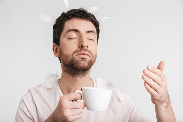 young man with bristle in casual t-shirt drinking coffee while standing under falling feathers isolated over white wall