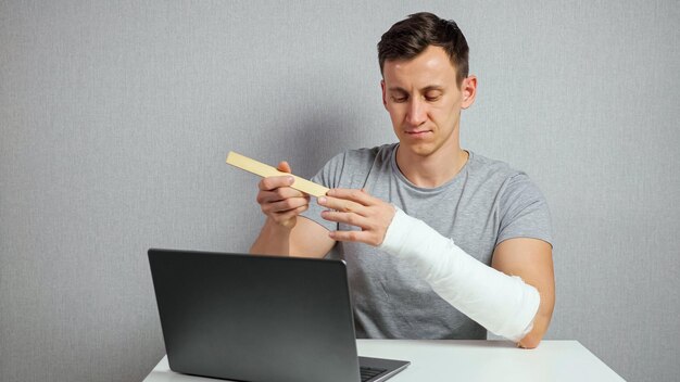 Young man with brunet hair in grey tshirt scratches injured forearm in gypsum band using wooden ruler and works on laptop sitting at desk