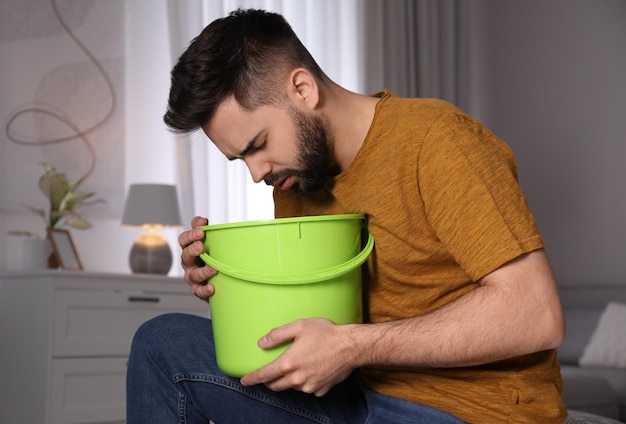 Young man with bucket suffering from nausea at home Food poisoning