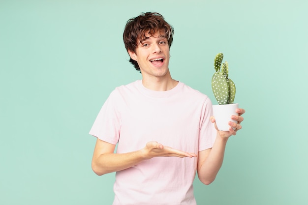 Young man with a cactus smiling cheerfully, feeling happy and showing a concept