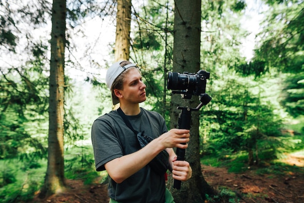 A young man with a camera on a stabilizer stands in the woods and creates travel content on a hike