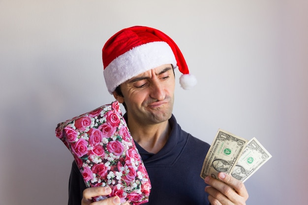 Young man with Christmas red hat holding wrapped gift and dollar bill with poor expression